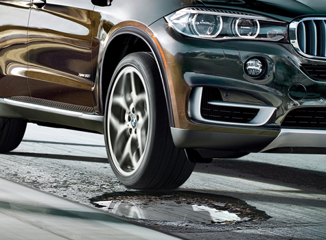 A close-up of the front of a brown BMW about to hit a large pothole on an asphalt road.