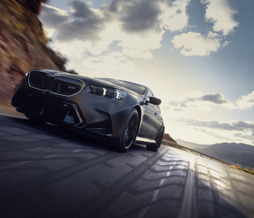 View looking up from the ground at a BMW M5 in motion on a road with mountains and blue sky and clouds in the background.