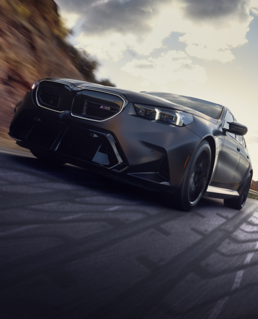 View looking up from the ground at a BMW M5 in motion on a road with mountains and blue sky and clouds in the background.